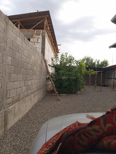 The image depicts an outdoor scene at a construction site. A tall, grey cinder block wall runs along the left side of the frame, with a concrete base. Beyond this wall, a partially constructed building is visible, with wooden roof beams and corrugated roofing material. A wooden ladder leans against the wall of the building, suggesting ongoing construction work.
In the middle ground, a lush green bush or small tree is growing. Behind the bush, there is a covered patio area with wooden posts supporting a simple roof. The ground is covered with gravel. The sky above is overcast, suggesting a cloudy day.
In the foreground, the hood and part of the roof of a silver car are visible, with a folded red and black patterned fabric draped over it. The setting appears to be a residential or semi-rural area in Жаны-Кыштак, Kyrgyzstan, given the construction activity and the style of the buildings and patio. The overall impression is one of a work in progress, with an overcast sky that doesn't provide strong cues about the time of day. There is no visible text in the image.