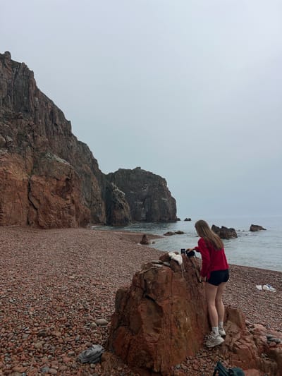 A woman in a red sweatshirt and black shorts kneels on a large rock at a beach on Russkiy Island, Russia, holding a camera and pointing it towards the ocean. She is wearing white sneakers and white socks. The beach is covered in small reddish-brown stones. Behind the woman are other rocks and the ocean. The sky is overcast.