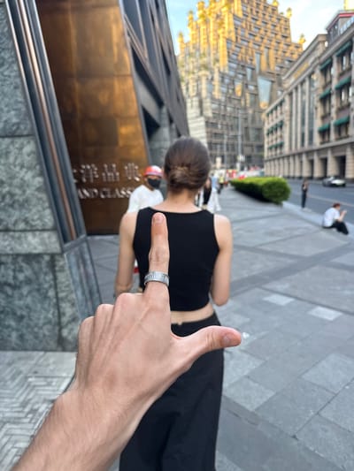 The image, oriented sideways, captures an urban scene from a first-person perspective. In the immediate foreground, a right hand is prominently displayed on the lower right, making a "thumbs up" gesture with the index finger extended and adorned with a distinctive silver-colored ring featuring a wavy or ridged texture.

In the mid-ground, slightly to the left and further back, the back of a person with dark hair (possibly in a bun) is visible, wearing a black sleeveless top and dark pants or a skirt. They are facing away from the camera, looking towards the background.

The background reveals a bustling city environment. Tall, modern buildings with varied architectural styles, including glass facades and light-colored exteriors, dominate the left side of the frame. One prominent building features a large, reflective golden-brown sign at its base. On this sign, partial Chinese characters are visible alongside the English words "AND CLASS." The ground is paved with large gray square tiles, suggesting a plaza or wide sidewalk. Further in the distance, blurred figures of other people are scattered, including one individual sitting on the ground wearing a white shirt and dark pants. Green shrubs or hedges line parts of the path.

The lighting suggests late afternoon or early evening, with a warm, golden glow illuminating parts of the buildings. The sky is bright but not directly visible. The overall scene depicts a casual public setting in Putuo District, China, conveying a sense of urban life and possibly a positive sentiment through the "thumbs up" gesture.