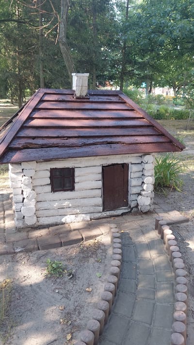 A model of a small, rustic log cabin is situated outdoors. The cabin has white-painted log walls, a dark brown wooden door, and a dark brown shingled roof. A white square chimney protrudes from the roof. A small, multi-paned window is visible on the left side of the cabin. The cabin is surrounded by a sandy area with scattered leaves and some sparse green plants. A paved walkway with a decorative border of small wooden posts curves away from the cabin. In the background, a line of trees, some coniferous and some deciduous, forms a backdrop. The setting appears to be a park or outdoor exhibition space, possibly within the city of Borodianka, Ukraine.