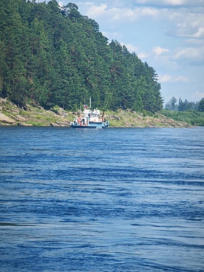 In a wide, blue river, a light-colored boat with a red and orange stripe along its hull is moored near a rocky, tree-covered embankment. The boat has a white superstructure with a Russian flag flying from a mast. Several people are visible on the deck of the boat. The embankment is densely covered with green coniferous trees, and large grey rocks are scattered along the shore. The water's surface is rippled, reflecting the blue sky and white clouds. The scene appears to be a natural landscape with a small vessel, likely in the city of Vysokogorskii, Russia, given the context.
