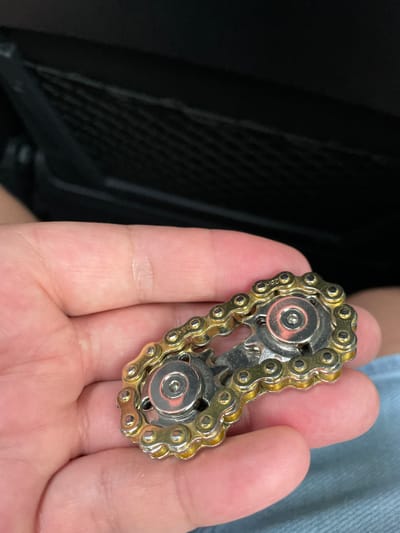 A person's hand is holding a fidget spinner made of a gold-colored chain and silver bearings. The fidget spinner is positioned in the center of the frame, with the hand occupying the lower left portion of the image. The background is out of focus and appears to be a dark, textured surface, possibly the interior of a vehicle. The lighting is soft and diffused, illuminating the fidget spinner and the hand.