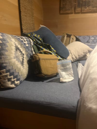 The scene presents a casual, indoor setting, likely a bedroom or living area. A bed is prominently featured, covered with blue bedding and several pillows. A brown backpack rests on the bed, partially obscured by a plastic bag labeled "Reynolds". A decorative patterned blanket or quilt is draped over the bed, adding visual interest.

The lighting is subdued, casting shadows and creating a warm ambiance. The location in the city of Livingstone, Zambia, cannot be definitively determined from the visual data alone. The presence of bedding, personal items, and the overall domestic atmosphere suggest a residential or lodging environment, potentially a hotel or private home. There are no people explicitly depicted in the scene.