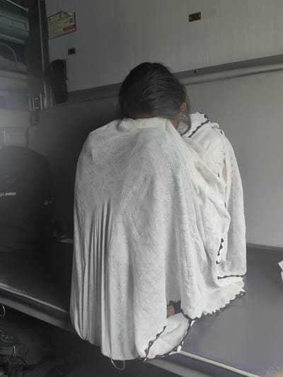 The image shows a person from behind, seated on a bench inside what appears to be a train compartment. The person is covered by a white cloth with a leaf-like pattern. The background suggests an interior space with a wall and a metal railing. The lighting is dim, casting shadows on the scene. The location within the city of Nepanagar, India, cannot be determined from the visual information provided in the image.