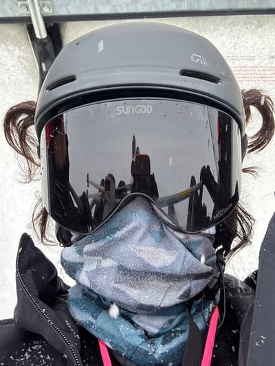 A close-up, frontal shot depicts an individual on a ski lift in St. Christoph, Austria, during active snowfall. The subject wears a dark grey Giro ski helmet and mirrored Sungod 4KO SNOW goggles, which reflect parts of a metallic ski lift structure and a red mobile phone held by an unseen individual, likely the photographer. Dark brown hair is visible exiting the helmet in two upward-curling sections on either side. A multi-colored (blue, grey, black camouflage pattern) neck gaiter covers the lower face and neck. The individual is dressed in a black winter jacket, with a neon pink strap visible on the right side. Numerous small snowflakes are visible on the helmet, goggles, neck gaiter, and jacket, indicating precipitation. A metallic beam is visible in the upper background, consistent with ski lift infrastructure, against an indistinct, snowy environment.