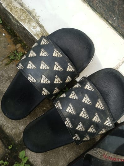 The image shows a pair of black Adidas slides, likely worn, sitting outdoors on a gray and white surface. Each slide features a thick strap across the top of the foot, patterned with repeated white Adidas logos and the brand name "Adidas" on a black background. They are somewhat dirty, hinting at regular use. The background suggests a partially shaded outdoor environment. Another pair of shoes is partially visible.