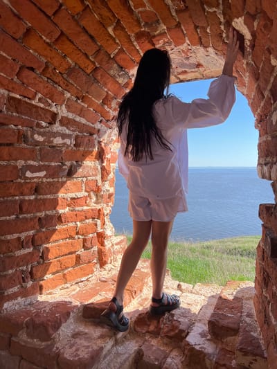 A person is seen within a brick archway, potentially a window or opening in a wall. The person is wearing a white shirt or dress and dark sandals. The person is holding onto the brick structure with one hand. The background visible through the archway includes water and grass. The location is indicated as Шуран, Russia.