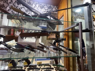 This image depicts the interior of a retail display case, likely within a store in Ayamonte, Spain, showcasing a variety of weapons. The case features multiple glass shelves supported by metal uprights, with a camouflage-patterned background on the upper section.
On the top shelf, a dark long gun, possibly an air rifle identified as "PREDATOR EX" and labeled "CALIBRE 4.5 M.M.," is displayed horizontally alongside a fixed-blade knife with a tan handle.
The middle shelf presents a wooden-stocked air rifle, marked "CALIBRE 5.5 M.M.," resting on white stands. Below it, an assortment of folding knives is visible, including one with an iridescent blade, another with an elaborately carved handle, and a balisong. A black revolver is positioned to the right. A folding knife with a blue and white patterned handle, bearing the text "POLICIA LOCAL," is clipped to a vertical support on the far right.
The bottom shelf contains several dark-colored handguns and numerous folding knives, featuring various handle designs such as camouflage and metallic finishes.
Reflections on the glass display case reveal elements of the store's interior, including a door or window in the background with a sign partially visible, stating "CONECTA 24 HORAS" and a telephone number (902 280 085). No individuals are present in the scene.