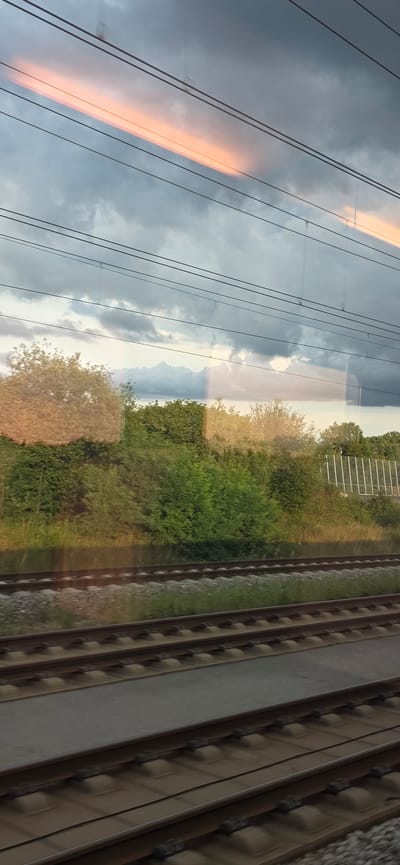 This media file depicts a reflection in a train window, capturing both the interior and exterior of a moving train. The dominant subjects are the train tracks, which run parallel to the right side of the frame, and the sky, which appears cloudy and features a prominent streak of orange light, possibly from a sunset or sunrise. Various trees and vegetation are visible along the side of the tracks, creating a green and autumnal landscape. The reflection also shows the interior of the train, including what appear to be overhead compartments or ceiling panels.
The activity taking place is travel by train. The sense of motion is conveyed by the blurred appearance of the landscape outside and the slight motion blur on the train tracks themselves. The time of day appears to be either early morning or late afternoon, given the warm, sunset/sunrise glow in the sky. The weather seems to be mixed, with dark clouds present alongside the brighter patches. There are no discernible people or text within the image. The overall mood is one of peaceful observation of the passing scenery during a journey. The location context of Allersberg, Germany, suggests a European railway environment.