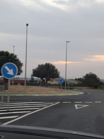 A roundabout is depicted with multiple signs indicating the direction of travel. A large blue sign with a white arrow pointing left is visible, along with smaller, circular blue signs showing the roundabout pattern. Striped pedestrian crossings are painted on the asphalt. Several streetlights line the roundabout. Trees are planted within the central island, adding a touch of greenery. The sky is overcast, suggesting it could be either early morning or late afternoon.