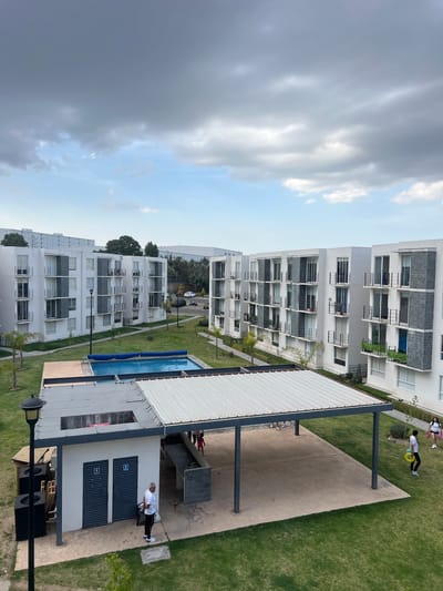 The image depicts an aerial view of a residential complex located in Sanctorum, Mexico. Several multi-story white apartment buildings with dark gray accents line a landscaped area with grass and trees. A swimming pool is visible adjacent to a covered recreation area with restrooms and picnic tables. People are present; one individual is near the restrooms, and two others are on the grass near the apartment buildings, engaged in a recreational activity using a yellow disc. The sky is partly cloudy, with a dark cloud covering the left side of the image.