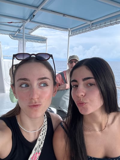 The image displays three individuals aboard a boat in West Bay, Cayman Islands. In the foreground, two young adult females are visible. The female on the left has brown hair pulled back, wears a black tank top, and a white beaded necklace; dark-framed sunglasses are perched on her head. Her lips are puckered, and her eyes are directed towards her left. The female on the right has long dark hair, wears a dark patterned top, and a thin gold chain necklace; her lips are also puckered, and her gaze is directed forward. An older adult male is situated in the background, between and behind the two females. He wears a green baseball cap, eyeglasses, and a light-colored shirt. A light-colored shoulder bag with a black bird logo is visible over his shoulder. The boat features white deck surfaces, structural posts, and a light blue canopy. Beyond the boat, dark blue open water is visible, extending to a horizon under a partly cloudy sky.