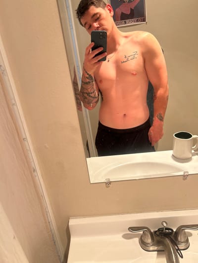 A man is taking a selfie in a bathroom mirror. He is shirtless and wearing black shorts. He has tattoos on his arms and chest. The bathroom has a beige wall, a white sink, and a vintage faucet. There is a mug next to the sink. A framed print hangs on the wall behind the man. The location appears to be a private residence. There are no indications that the photo was taken in Reno, Nevada.