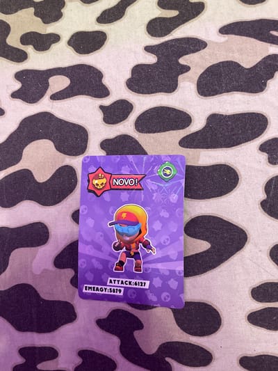 The image shows a trading card with a cartoon character on a purple background. The card has the text "NOVO!" and "ATTACK:6127" and "EMEAGY:5879". The card is lying on a surface with a camouflage pattern. There is no information about the city of Vaganovo Massiv, Russia, in the image.