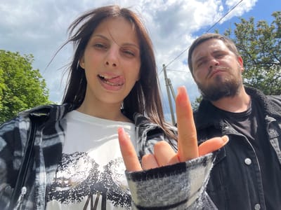 Two people, one male and one female, are in the frame. The image appears to be taken from a low angle, looking upward toward the subjects. The woman is sticking out her tongue. The man has a beard. The woman is making the "rock on" sign with her hand. They are outside in an area with trees and cloudy skies. Given the prompt, the location is Blizhniy Khutor, Moldova.