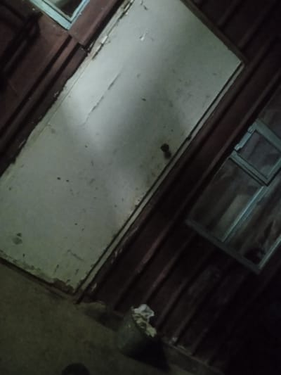 The image depicts a dimly lit, angled view of a roughly constructed building exterior. A large, pale, weathered door dominates the center of the frame, leaning slightly to the left. To the right of the door, a small, dark-framed window with multiple panes is visible. The building's exterior walls are made of horizontal wooden planks, colored a dark reddish-brown, with a slightly corrugated texture. Above the door, the top edge of another window or opening is partially visible, its frame a lighter color. In the lower right corner, a dark, cylindrical object, possibly a bin or container, is visible with some debris or trash on top. The ground in the foreground appears to be concrete or packed earth. The overall impression is one of an outdoor, utilitarian structure in a rural or semi-rural setting, likely in Taldı-Suu, Kyrgyzstan, given the context. The lighting suggests it might be nighttime or a very shaded area. There are no people visible in the image.