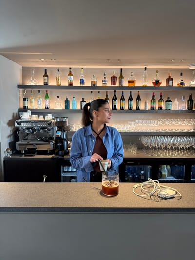 An interior scene depicts a female individual standing behind a dark gray speckled bar counter within what appears to be a bar or restaurant in Marche-lez-Écaussinnes, Belgium. The woman, with dark hair pulled into a ponytail and wearing a blue and white checkered shirt over a brown strap top, holds a small white cup with a black stirrer in her left hand, gazing to her right. Behind her, against a light gray wall, two horizontally illuminated shelves display an array of approximately 30-40 glass bottles containing various alcoholic beverages, including clear, amber, and dark liquids. A clear glass decanter is on the top shelf to the far left. To the woman's left, on a lower black counter, a professional stainless steel espresso machine and a black coffee bean grinder are situated alongside stacks of white ceramic cups. To her right, numerous rows of empty, clear stemmed glassware are meticulously arranged on a shelf. Below the counter, dark cabinetry and two glass-fronted refrigerated units with bottles visible inside are present. On the dark gray counter in the foreground, a clear glass filled with a dark beverage, containing several dark, round objects and a black straw, is positioned directly in front of the woman. To the right of the glass, a coiled white charging cable with a power adapter is visible. The overhead ceiling features recessed lighting.