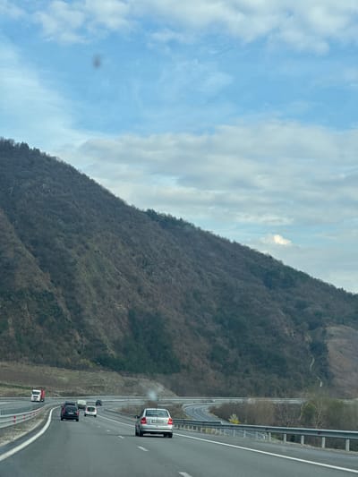 The image depicts a highway adjacent to a mountain, likely taken from a moving vehicle. The sky is partly cloudy. A mountain covered with trees in their dormant, leafless state dominates the left side of the frame. On the right is a multi-lane highway with several vehicles, including a silver car, a black SUV, and a truck. The location is identified as Jeleznitsa, Bulgaria, suggested by the terrain and road infrastructure. The angle of the photo implies it was taken from a higher position, inside a car, moving alongside the highway.