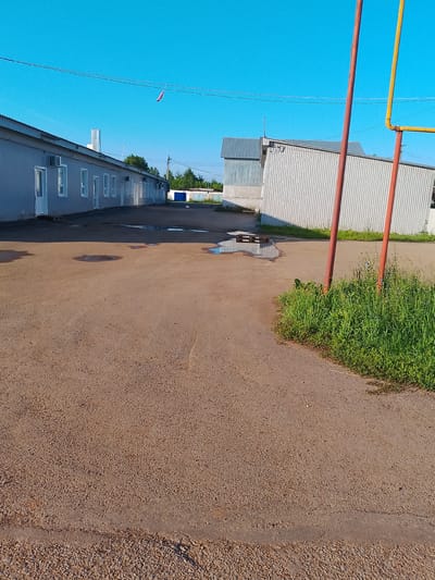 A bright, sunny day with a clear blue sky is depicted. In the foreground, a dirt and gravel road surface is visible, with a strip of rough asphalt at the bottom. To the right, a patch of tall green grass with a few small purple flowers grows. Two upright poles, one reddish-brown and the other yellow, stand in the grassy area. In the middle ground, a large, light gray, corrugated metal building stretches across the scene. It has a row of white-framed windows and doors along its length, and several outdoor air conditioning units are mounted on its side. A few puddles of water are present on the ground in front of this building. Further back, other corrugated metal structures are visible, including one with a peaked roof. Power lines are strung across the sky, with a small red and white object hanging from one of them. In the distance, trees and a blue building are seen. The overall scene appears to be an industrial or commercial area, possibly a facility or warehouse complex, under a summer sky. There are no people or animals visible in the image.