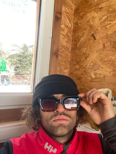 A male subject is depicted in a close-up, chest-up view, situated indoors within a structure in La Salle-les-Alpes, France. The interior walls are constructed from oriented strand board (OSB). The subject wears a black knit beanie and a red and black jacket with "HH" branding on the left chest. He has a beard and mustache. He wears large, square-framed sunglasses with a brown tortoise-shell pattern, which reflect a snowy outdoor scene. His right hand is raised, with fingers gripping the right temple of his sunglasses, indicating an adjustment. A gold-colored earring is present in his left earlobe. To his left, a white-framed window provides a view of a snow-covered outdoor landscape, featuring coniferous trees, distant mountainous terrain, and a red net barrier. A green graphic resembling a stylized tree is affixed to the lower window pane. Faint green writing, possibly "SWAP?", is visible on the window's interior surface. A partially visible cream-colored, possibly fur-lined object and a dark item are situated to the subject's right.