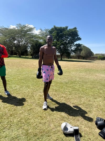 The image captures an outdoor scene in Olkaria ward, Kenya, under a clear blue sky with scattered white clouds. The setting is a large, manicured grassy field with green and some dry patches. In the foreground and center, a dark-skinned adult male stands facing the camera, smiling. He is shirtless, wearing pink shorts with a light blue jellyfish pattern, black boxing gloves on both hands, and grey athletic shoes. To his left, partially visible at the image's edge, is another dark-skinned adult male, wearing a red and black patterned long-sleeved shirt, green shorts, and grey athletic shoes. On the ground in the immediate foreground, a grey boxing glove with a black strap and a partially visible black boxing glove are present. The background features several large, green trees, including Acacia species, and a low, light-colored stone wall or fence stretching across the mid-ground. Strong sunlight casts distinct shadows of the individuals and objects onto the grass.