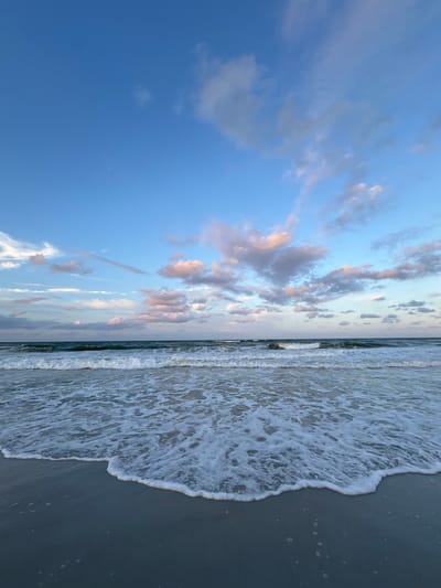This is a vertical, eye-level, medium shot of the ocean at Jacksonville Beach, Florida, United States of America. The ocean is on the right side of the image, with a sandy beach in the foreground. The sky is a clear blue with scattered clouds that have pink and white hues. The waves are breaking on the shore, with white foam washing onto the beach. The lighting suggests it is either sunrise or sunset. There are no people or man-made objects visible in the frame.