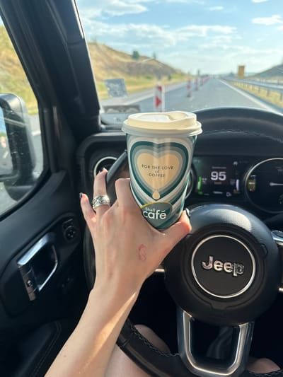 A photograph shows the interior of a Jeep vehicle, focusing on the driver's side. The driver's hand is holding a "Shell café" coffee cup with the slogan "FOR THE LOVE OF COFFEE." A ring is visible on the driver's finger, and a small, red tattoo is on their wrist. The Jeep's steering wheel, displaying the "Jeep" logo, is visible. The digital dashboard shows the vehicle traveling at 95 km/h. The exterior of the car shows a highway landscape under a bright, slightly cloudy sky. The photograph was taken on the A2 highway near Hadzhidimitrovo, Bulgaria, evidenced by signage and the overall landscape visible through the windshield.
