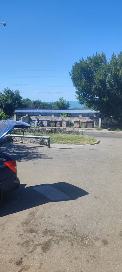The image depicts an outdoor scene on a clear, sunny day, likely in the city of Svitlovodsk, Ukraine, given the context. In the foreground, the rear of a dark blue car with its trunk open is visible on an asphalt surface. The asphalt appears somewhat worn and has some dark stains. A prominent shadow of the car is cast on the ground. Behind the car, a low, decorative stone wall with a metal fence separates the paved area from a patch of green grass.
Further back, a line of large, weathered metal dumpsters stands against a brick wall topped with a corrugated metal roof. One person, dressed in a blue shirt and dark pants, is seen walking near the dumpsters, carrying a white bag. Beyond the dumpsters, a line of trees is visible, and further in the distance, the blue expanse of a body of water, possibly the Dnieper River or the Kremenchuk Reservoir, is visible under a clear blue sky. Overhead, thin power lines are strung across the sky. The overall impression is a utilitarian area, likely a collection point for waste, situated in a location with a natural backdrop.