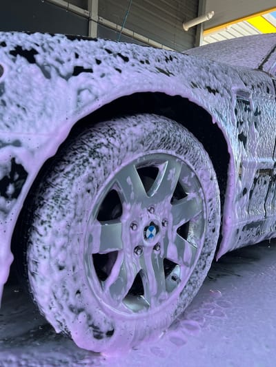 A close-up shot focuses on the front passenger side wheel and fender of a dark-colored BMW, which is covered in pink foam, indicating it is being washed. The car is positioned in what appears to be an outdoor car wash facility. The wheel, a silver alloy with multiple spokes, features the BMW logo at its center. The tire is also heavily coated in foam. The fender arch and the lower portion of the car's door are visible, also obscured by the pink foam. The background shows the corrugated metal siding of a structure and yellow-colored roofing. The ground beneath the car is wet and covered in a layer of the same pink foam. The scene is well-lit by natural light. The location is identified as Velyki Hai, Ukraine.