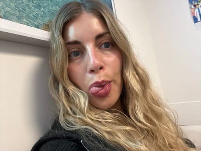 A close-up selfie features a young woman looking directly at the camera with a playful expression, her tongue slightly extended from her mouth. She has long, wavy blonde hair that frames her face and falls over her shoulders, and distinct blue eyes. Her lips are adorned with a dark, reddish-purple color. Her skin exhibits a natural, dewy sheen. She is dressed in a dark, textured sweater.
The background elements suggest an indoor setting. To her left, a white shelf is visible, above which a light blue, textured surface, possibly a wall covering or artwork with a wave-like pattern, can be seen. To her right, a plain white wall extends, with a small portion of a colorful illustration or poster featuring cartoon-like figures just barely visible in the upper right corner. The overall lighting indicates it is daytime indoors. The photograph was taken in an indoor environment, likely a room or office, in Les Lilas, France. No discernible text is present in the image.