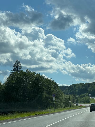 The image captures a scenic view from a moving vehicle on a sunny day in Kunya, Russia. The perspective is from the passenger side, looking outwards. The scene is dominated by a bright blue sky filled with dramatic, fluffy white and gray clouds. Below the sky, a dense, lush green forest lines the right side of the frame, with trees forming a thick canopy that extends towards the horizon. A paved road, separated by a white line, runs alongside the forest, suggesting the vehicle is traveling on a highway or a main road.

In the foreground, a dark-colored vehicle, possibly an SUV, is partially visible on the right side of the image, indicating the point of view is from inside another car. A single traffic sign is visible on a pole along the roadside, featuring a blue circle with a red border and a diagonal red line through it, signifying a prohibition. Below this, there's another sign with a blue upward arrow and the text "1.0 km". The overall atmosphere is one of a pleasant summer day, with clear weather and good visibility. The time of day appears to be late morning or early afternoon, given the bright sunlight casting shadows and illuminating the clouds. The location, Kunya, Russia, adds a geographical context to the picturesque natural scenery.