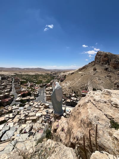 The image depicts a view overlooking the city of Ma'loula, Syria, from a high vantage point on a rocky cliff. In the foreground, the edge of the cliff is visible with sparse vegetation. The town below is densely packed with buildings, with a network of streets and roads visible. In the distance, the landscape transitions into arid hills under a clear blue sky with scattered clouds. A large, draped object is positioned at the immediate edge of the cliff.