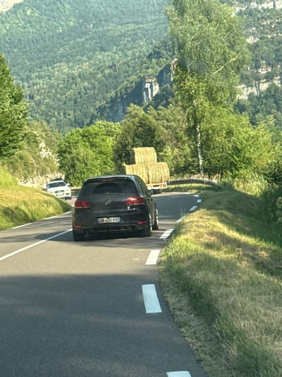 In this vertically oriented image, a dark-colored Volkswagen Golf is captured in motion on a paved road winding through a lush, green mountainous landscape. The car is positioned on the right side of the frame, its rear facing the viewer. The road features white dashed lines on the right edge, indicating a lane marker. To the left of the road, a steep grassy embankment rises, with several large, rectangular hay bales stacked on a yellow trailer. Beyond the embankment, a dense forest covers the hillside, with trees of varying shades of green creating a textured canopy. The terrain suggests a rural or mountainous setting, consistent with the location context of Lavans-lès-Saint-Claude, France.
Further down the road, a white SUV is visible, also in motion. The overall scene evokes a sense of travel and exploration through scenic countryside. The lighting suggests it is daytime, with sunlight casting shadows on the grass and road, and highlighting the textures of the foliage. No people are directly visible in the image, but the presence of vehicles implies human activity.
The license plate of the Volkswagen Golf is partially visible and reads "CM 603 RY". The image has a slightly grainy quality, possibly due to the capture or processing of the photograph. The time of day appears to be afternoon, given the angle of the shadows.