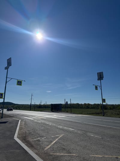 The image depicts a pedestrian crosswalk in Krilovo, Russia. The crosswalk is marked with white stripes on a grey asphalt road, with pedestrian crossing signs on posts with solar panels. The sky is blue and clear, with the sun visible. Power lines run parallel to the road in the background. A line of trees is visible beyond the road.
