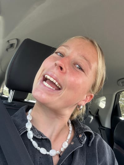 A young blonde woman in a car smiles broadly at the camera. She wears a black denim jacket and a white pearl necklace. A seatbelt crosses her chest. The car's interior is visible in the background, including the headrest and ceiling. She also has shiny silver hoop earrings.