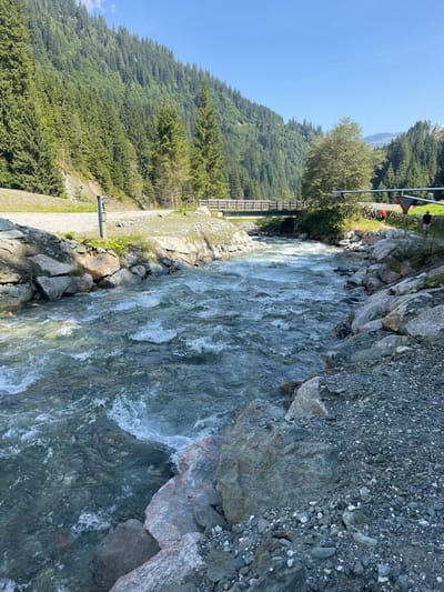 The image displays a clear, fast-flowing mountain river traversing a forested valley in Reitlehen, Austria, under a cloudless blue sky. The river occupies the central and foreground portions of the scene, exhibiting turbulent, greenish-blue water with visible rapids. Its banks are unpaved and composed of large boulders and smaller gravel. Dense conifer forests cover the steep mountain slopes on both sides of the valley.

On the left riverbank, a gravel path runs parallel to the water, where a dark, vertical post with a horizontal protrusion is present in the foreground. In the mid-ground, a wooden-decked bridge with green metal supports spans the river. On the right riverbank, near the bridge, a group of approximately three to four indistinct individuals are visible, and a single individual wearing a dark hat is partially visible walking away from the camera. A large, light-colored metal structure, possibly a utility pole or support, with an attached triangular warning sign, is partially visible on the far right of the frame.
