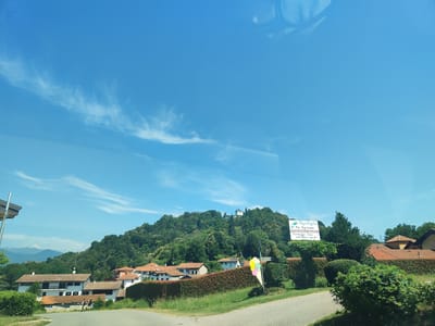 This is a wide-angle photograph taken on a sunny day from a vehicle, likely in Borghetto, Italy. The scene captures a rural landscape with a clear blue sky dotted with wispy white clouds. In the foreground, a paved road curves gently to the right. To the left of the road, a white building with a wooden roof overhang is partially visible, suggesting it is part of a structure or perhaps a bus stop.

Further back, a cluster of buildings with red-tiled roofs and white walls are nestled amongst lush green trees and hedges. The buildings appear to be residential homes. Behind the houses, a densely forested hillside rises, with a prominent white building, possibly a church or chapel, situated on its crest.

To the right of the frame, a signpost stands with an illustration of a colorful ice cream cone. Adjacent to this, a white sign with green text reads "L'Agrifoglio Az. Agricola" and below that, "Produzione e Vendita Formaggio Latte e derivati" (Production and Sale of Cheese, Milk, and derivatives). A small arrow points to the right on the sign. Two people, a man and a woman, are walking on a path in front of the houses. The overall atmosphere is peaceful and idyllic, characteristic of the Italian countryside.