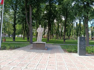 The image depicts a monument to Józef Piłsudski located in Grójec, Poland. The monument consists of a stone statue of Piłsudski in military attire, standing on a rectangular pedestal inscribed with his name. To the right of the Piłsudski monument is a tall, dark stone slab with Polish text and emblems. The monuments are situated in a park-like setting with a paved walkway made of square stones in the foreground. Lush green grass and numerous tall trees with dense foliage surround the monuments. A white flagpole with a red and white flag is visible to the left of the frame. In the background, there are more trees, a paved road, and glimpses of buildings and playground equipment, suggesting it is an urban park. The scene is illuminated by natural daylight, casting soft shadows.