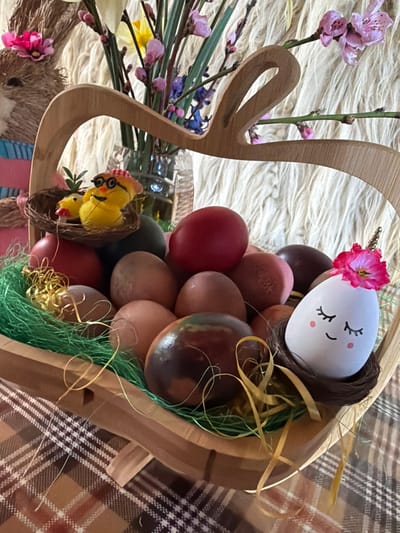 A close-up shot shows an Easter basket filled with colored eggs and decorative items, likely taken indoors, perhaps in a household setting. The basket is made of light-colored wood, possibly in the shape of a rabbit. Inside the basket are several Easter eggs, various shades of brown, red, and other colors. One egg is decorated as a unicorn with a flower attached. Artificial green grass and gold ribbon are also placed within the basket, contributing to the Easter theme. A small nest with two yellow chicks is placed in the basket as well. The background consists of a white, fluffy surface and a brown plaid surface. A vase with flowers and a bunny decoration can be seen in the background. The image does not provide information about the location in the city of Davidkovo, Bulgaria.