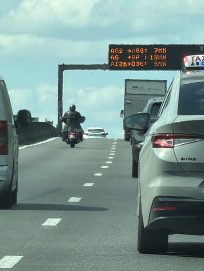 This media file captures a traffic scene from the perspective of a vehicle. The primary focus is on a digital road sign displaying traffic information. The sign, oriented vertically, shows several lines of text in orange luminescence against a black background. The text includes "TAX", "7MN", "AGB", ">BP", "15MN", "SMN", and "A126+D36:".
The scene also includes a white SUV with "ENYAQ" visible on its rear. A truck is partially visible to the left of the road sign. Further down the road, a motorcycle with a rider wearing a helmet is seen in traffic. White diamond-shaped markers are present on the road, indicating lane dividers. The sky is visible in the background, appearing overcast or hazy. The overall setting is a highway or a busy road, suggesting a traffic jam or slow-moving traffic. The time of day is not explicitly clear, but the lighting suggests it is daytime. There are no strong emotional cues conveyed. The location context provided is Savigny-sur-Orge, France.