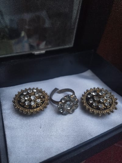 The image depicts three pieces of costume jewelry laid out within a dark-colored box with a reflective lid. The scene is located in Gerasimovskoye, Kazakhstan. The jewelry consists of one adjustable ring and two circular items, possibly brooches or earrings, all featuring a central cluster of multiple clear, faceted stones set in an aged, ornate metallic base, likely brass or a tarnished gold-tone alloy. The ring is centrally positioned, with the two circular items flanking it. All items rest on a light-colored, possibly white or off-white, fabric lining the bottom of the box. The reflective surface of the open lid shows a blurred, indistinct reflection of the surrounding environment. No individuals or active interactions are visible in the scene.