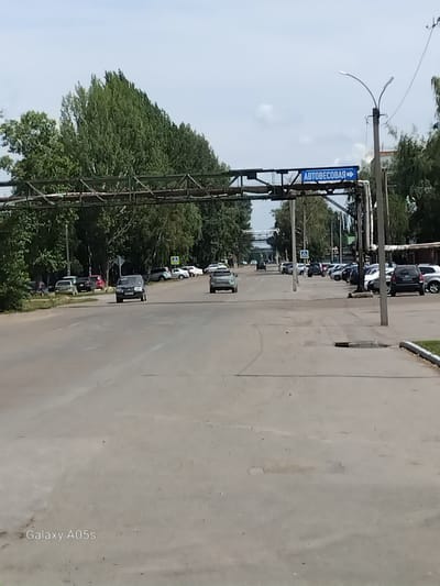The image shows a street scene in the city of Aleysk, Russia, under a cloudy sky. In the foreground, a wide asphalt road leads into the distance, with parked cars visible on the right side. A blue sign with white lettering, reading "АВТОБЕСОВАЯ" (Autobeskova), and an arrow pointing to the right, is suspended over the road from a metal structure. A lamppost stands on the right side of the road. Trees line the left side of the street. In the background, more trees and buildings are visible. The overall scene depicts a typical urban environment on a summer day.