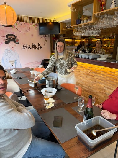 This image captures an indoor dining scene at an Asian-themed restaurant in Walferdange, Luxembourg.
In the foreground, a light-skinned man with dark hair, wearing a light grey sweater and blue jeans, is seated on the left, looking directly towards the viewer with a slight open-mouthed expression. His arms are crossed. To his right, a light-skinned woman with blonde hair and glasses, wearing a cream floral-patterned turtleneck sweater and white pants, stands leaning over a dark wooden table. She is holding small white food items, appearing to place one into a bowl. A hot pot apparatus is centrally located on the table, surrounded by various dishes, including a white bowl of green leafy items and small plastic containers of white food items. A glass of Battin beer, several empty champagne flutes, a bottle of Charles Montaigne Brut champagne, and a clear plastic container with a dark sauce and a ladle are also on the table. A black mobile phone displaying "15:51" rests on a grey placemat. Part of another diner in a red top is visible on the far right.
The background features a large mural on the left wall depicting a geisha with cherry blossoms and the Japanese characters "醉美櫻花" (Zui Mei Ying Hua). A large, round, light-colored lantern hangs from the ceiling. A flat-screen television is mounted on the wall above the mural. On the right, a wooden bar counter with illuminated horizontal slats is visible, behind which a light-skinned woman with dark hair, wearing a leopard-print top, stands, looking towards the left. Shelving above the bar holds rows of inverted wine glasses, decorative boats, and small plants. The restaurant has a modern, brightly lit interior with a distinct Asian aesthetic.