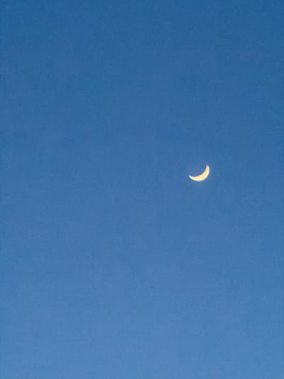 The image displays a clear, consistent mid-blue sky, observed from Connah's Quay, United Kingdom. Positioned in the upper-right quadrant of the frame is a distinctly illuminated crescent moon, exhibiting a luminous yellow-gold coloration. No other celestial bodies, atmospheric phenomena, terrestrial objects, structures, individuals, or discernible actions or interactions are present within the frame. The environment is solely atmospheric, depicting the sky and a singular celestial object.
