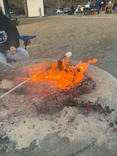 In the city of Old Malibu, United States of America, a close-up shot focuses on a bonfire pit with a marshmallow being toasted over the flames. The bonfire is constructed from a large log within a circular concrete structure. Intense orange flames erupt from the burning wood, casting a warm glow. A single white marshmallow is skewered on a stick, positioned directly above the fire, appearing to be in the process of toasting. Two other sticks with what appear to be marshmallows or other food items are also present, one held by a person partially visible in the frame. The person is wearing a black hoodie with white and blue graphics and grey sweatpants, sitting in a blue camping chair. The surrounding environment is sandy and appears to be an outdoor recreational area, possibly a campsite, with dry grass and a few distant structures and vehicles visible in the background, suggesting a casual gathering.