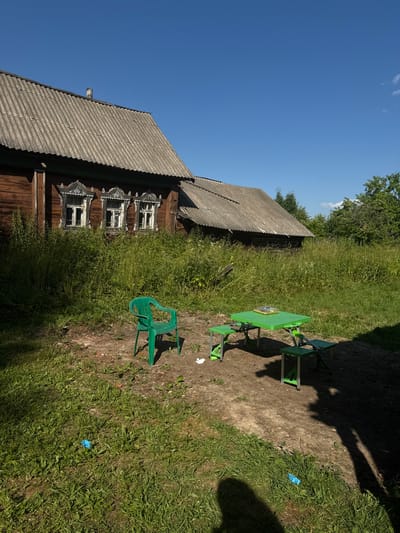This media file captures an outdoor scene on a sunny day in Ivan'kovo, Russia. The primary subjects include a weathered wooden house with ornate window frames, a bright green picnic table with attached benches, and a single green plastic chair. The house has a corrugated roof and is surrounded by tall, green grass and some dirt pathways. Trees are visible in the background, and the sky is a clear, vibrant blue with a few wispy clouds.
The scene appears to be set up for outdoor leisure, with the picnic table and chair suggesting a place for relaxation or dining. The overall impression is one of a rural or semi-rural setting, likely during the daytime, given the strong sunlight and shadows cast by the objects and foliage. There is no discernible activity taking place; the scene is static, presenting a snapshot of the environment.
Notable details include the intricate carvings on the window frames of the house, hinting at traditional Russian architecture. The green plastic furniture provides a modern contrast to the older, rustic building. A small blue object, possibly trash, is visible on the grass near the pathway. The time of day is clearly indicated by the long shadows, suggesting it might be late morning or early afternoon. The weather is pleasant, with bright sunshine. There is no visible text in the image.