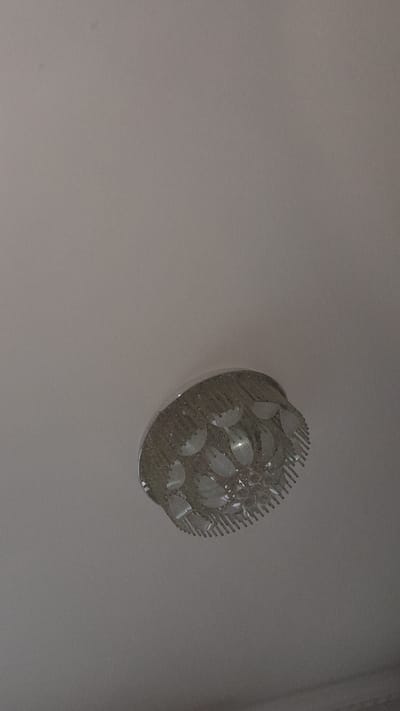 An ornate chandelier hangs from a plain, light-colored ceiling. The chandelier is round and appears to be made of glass or crystal elements that are arranged in a floral or petal-like pattern around a central metallic fixture. The glass elements are translucent and have a ribbed texture, catching the light. The ceiling is smooth and unadorned, providing a neutral background for the decorative light fixture. The image is shot from a low angle, looking upwards at the chandelier. There are no people or other objects visible in the frame. The scene appears to be an indoor setting, likely a room in a residential or commercial building. The location in the city of Ismoili Somoni, Tajikistan cannot be determined from the visual information provided in the image.