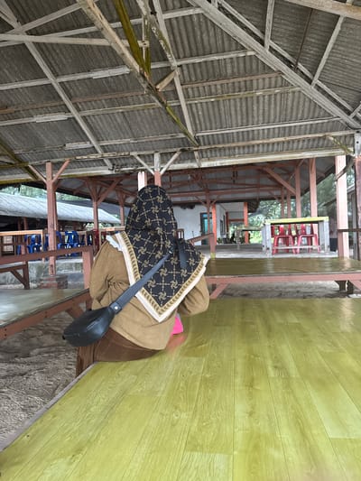 In Meunasahbale, Indonesia, a person wearing a brown hijab with a gold pattern and a brown outfit sits at a light green wooden table under a weathered, open-air structure. The structure has a corrugated metal roof supported by wooden beams. Behind the person, several red chairs are visible around a table. The ground appears to be sandy.