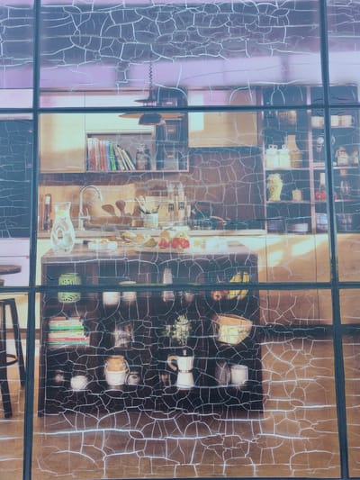 This image depicts a store window display in Bielany Wrocławskie, Poland, featuring a simulated kitchen scene. The display is divided by dark vertical and horizontal lines, resembling panes of glass. The "kitchen" shows a countertop with a sink, faucet, and various items such as a glass pitcher of water, bowls of fruit, and loaves of bread. Behind the counter, shelves are filled with books, jars, and kitchenware. Below the counter, more shelves display decorative items, mugs, and a coffee maker. The overall effect is a stylized representation of a kitchen, likely intended to attract customers to the store. The display appears to be either printed on or applied to the window itself.