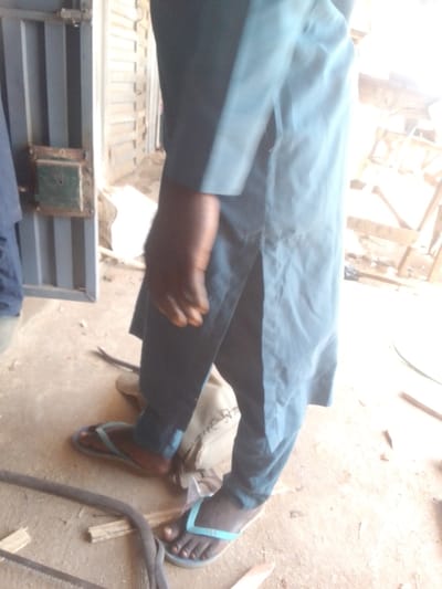 This image depicts a segment of an indoor or semi-sheltered area, potentially a workshop or storage space, in Nigeria. The precise city location within Nigeria cannot be determined from the visual information provided.
The scene features at least two individuals. In the foreground, a person is standing, captured from mid-thigh to the feet, and including part of their right arm and hand. This individual wears a dark blue or teal loose-fitting tunic or shirt with a side slit on the left, paired with matching trousers. Light blue/teal flip-flops are worn on both feet; the right flip-flop bears the text "SLS". The visible skin is dark brown. To the far left, a second person's dark blue garment and a light-colored shoe or boot are partially visible on the ground.
The floor is a light brown, uneven surface, appearing to be concrete or packed earth, covered with dust and debris, including wood shavings and small pieces of wood. A dark, coiled object, possibly a hose, cable, or flexible saw blade, lies on the floor near the foreground individual's feet. A light-colored, possibly canvas bag with dark markings rests on the ground behind the main individual's legs.
To the left, a grey metal gate or door with vertical bars is visible. Attached to it is a rectangular, green-painted metal lock housing, exhibiting signs of wear and rust. The background to the right features blurred, light-colored wooden structures, suggesting a raw material storage area or unfinished carpentry. The scene is brightly lit, likely by natural light from an unseen source to the right, casting areas into strong light and shadow.